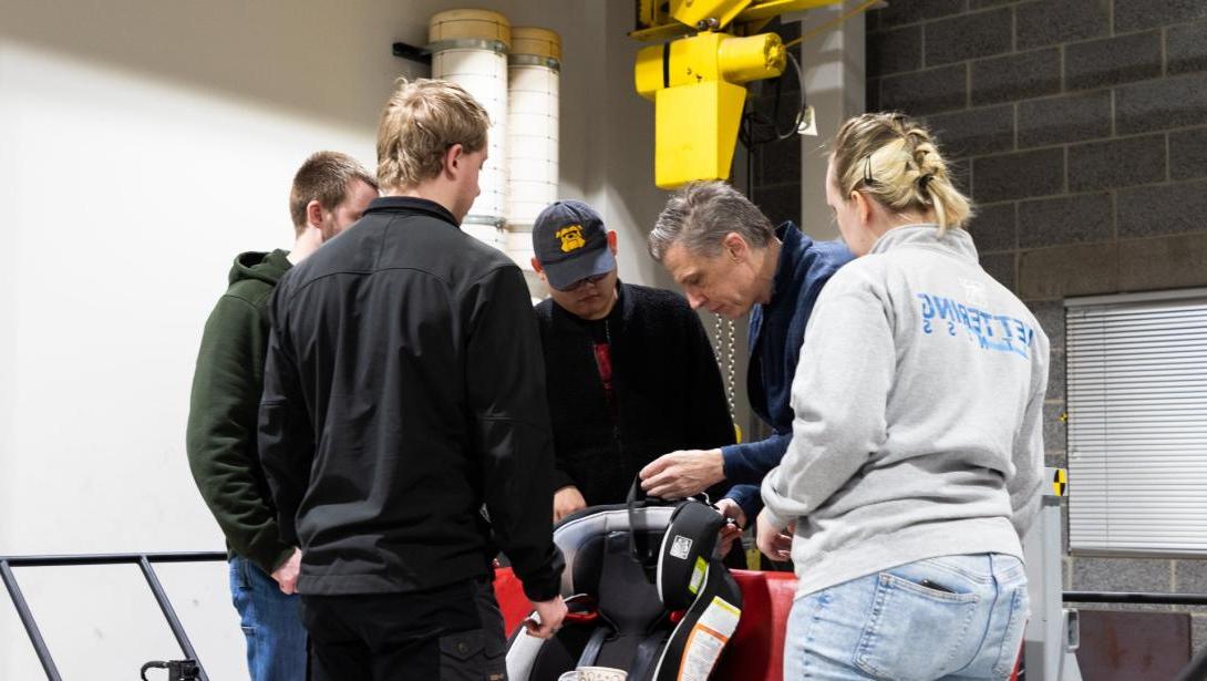 A group of students watch a professor work with a car seat in the Crash Lab