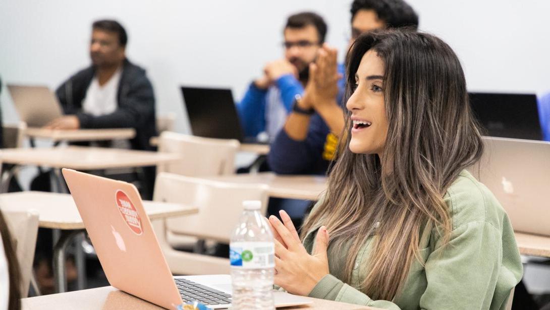 A student sits at a desk with an open laptop in a classroom with other students in background