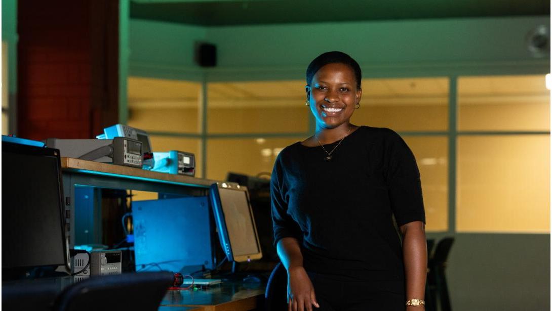 An individual in business attire poses in front of a desk with equipment